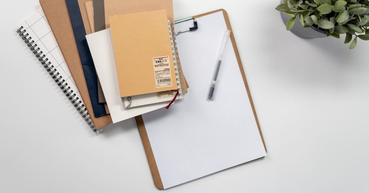 A bird's eye view shows us a clipboard holding a blank A4 piece of paper, sitting in the centre of the image, on a white table. On top of the clipboard sits a small pile of brown-coloured envelopes and spiral-bound notebooks. To the top right corner of the image is a potted plant.