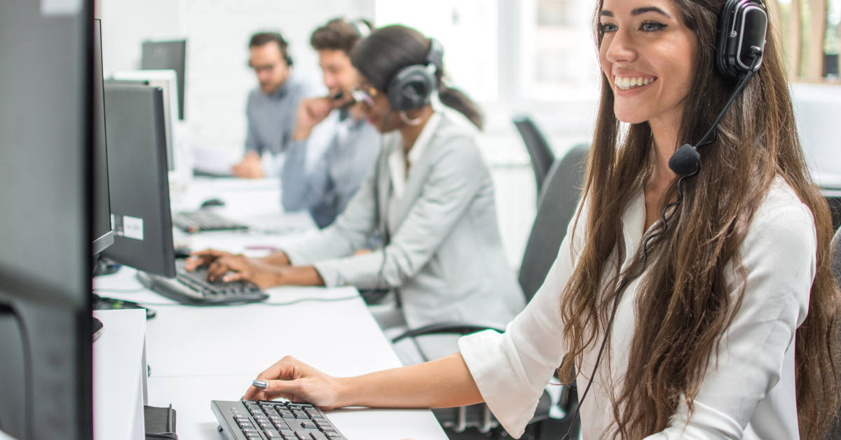 In the foreground sits a young women sat at a computer. She wears a headset and smiles at her computer screen. In the background are other workers in headsets.