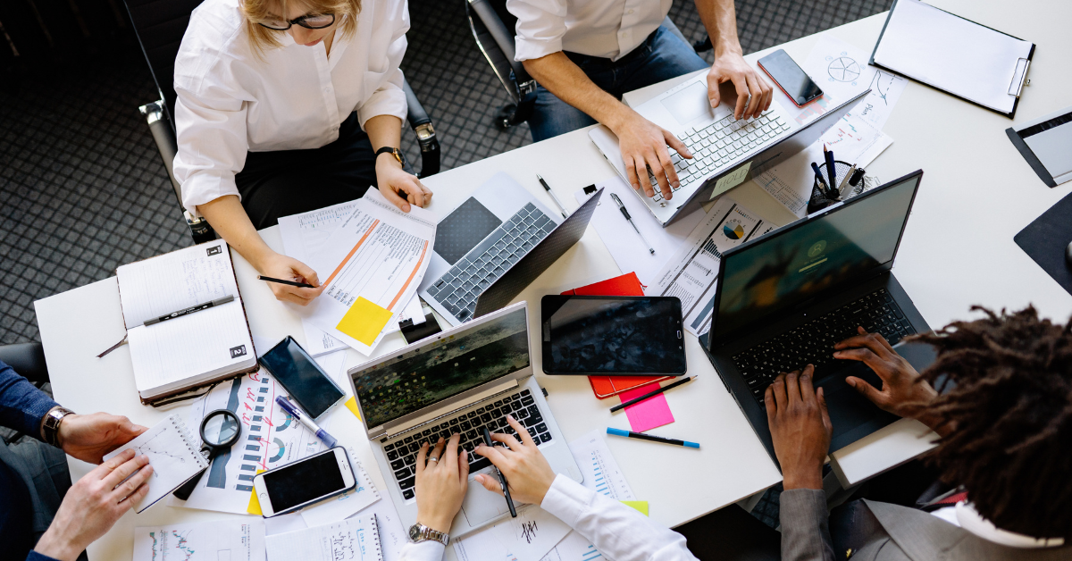 Bird's eye view of a large office table with five people sat around it. On top of the table are laptops, notebooks, post-it notes and clipboards.