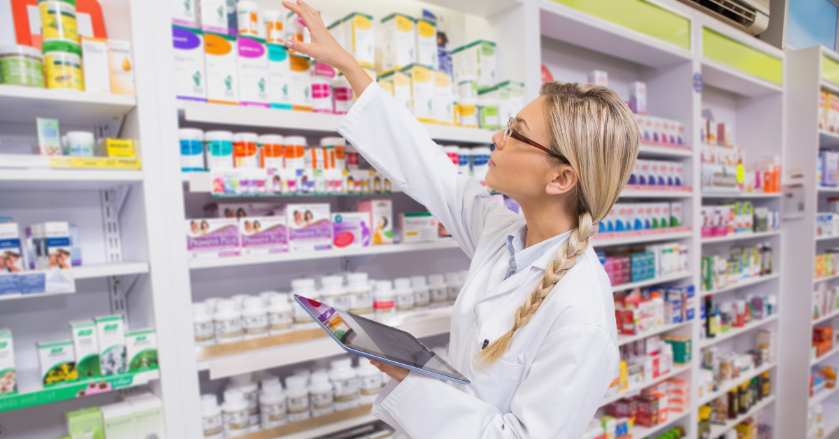 In the centre of the image, we see a young woman in a white lab coat reaching for something from the stacked shelves behind her. The shelves display various medicines and bottles of tablets.