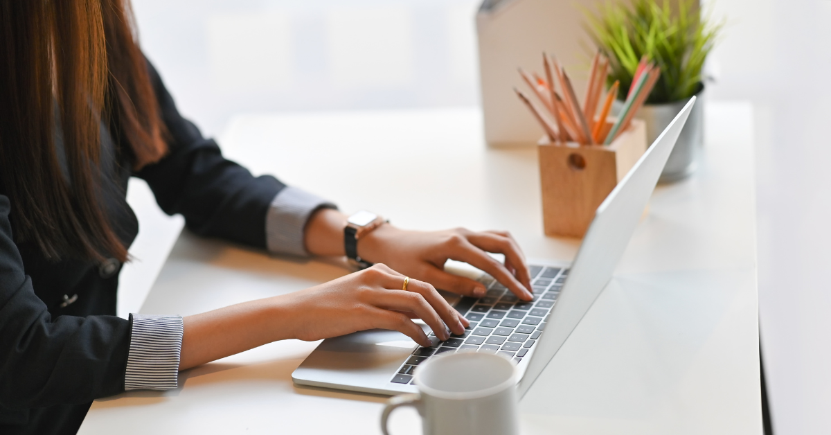 A pair of hands type at a laptop keyboard. In the foreground is an empty mug and in the background is a pot filled with pencils.