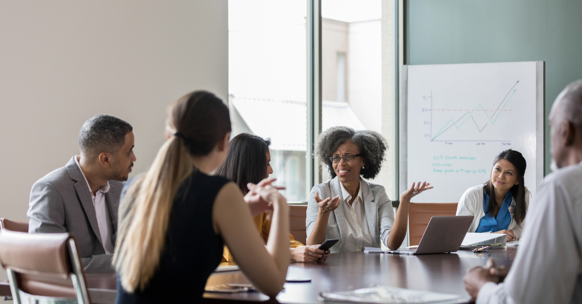 A group of six people sit around a table in a meeting, by a large window. In the background is a whiteboard with some writing on it. The focus of the image is a black woman in the centre, who is talking animatedly to the people to her right.