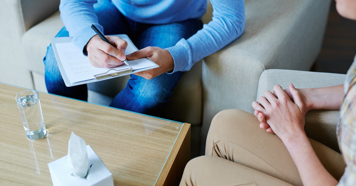 Image shows the torsos and legs of two people, each sat in an armchair at the corners of a wooden table. The person in the background is writing on a clipboard, while the person in the foreground speaks to them. On the table is a full glass of water and a box of tissues.