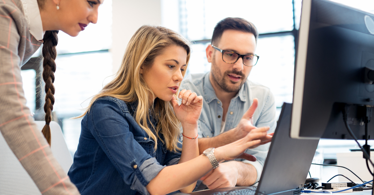 Three people surround a laptop. A blond-haired woman sits in the centre, with a man to her left and a woman standing over her right shoulder. They are talking about what they can see on the laptop screen.