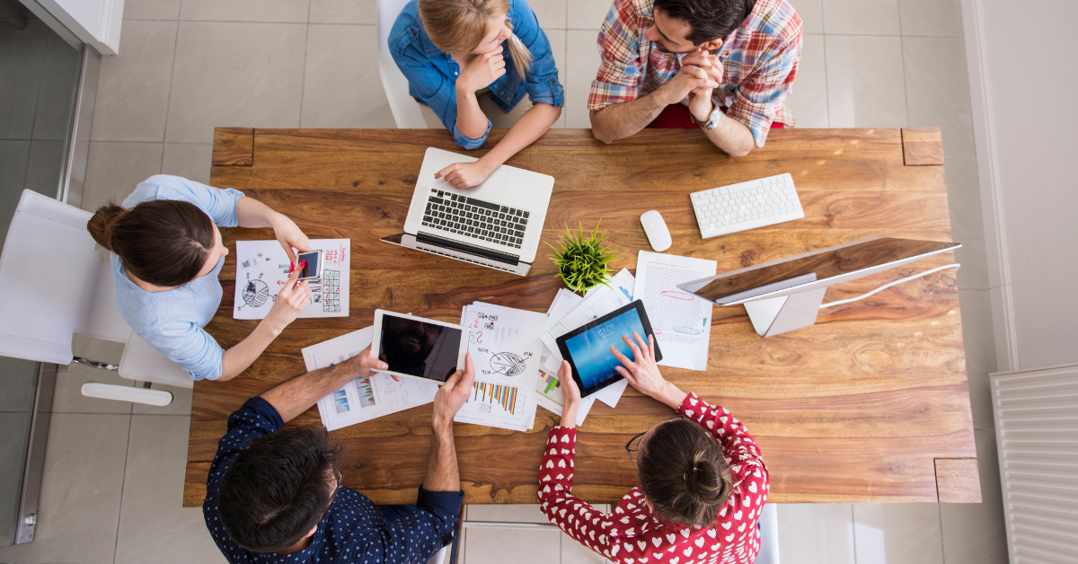 From a bird's eye view we see five people sat around a wooden table. In front of them are laptops and tablets, and sheets of paper displaying data in various formats.