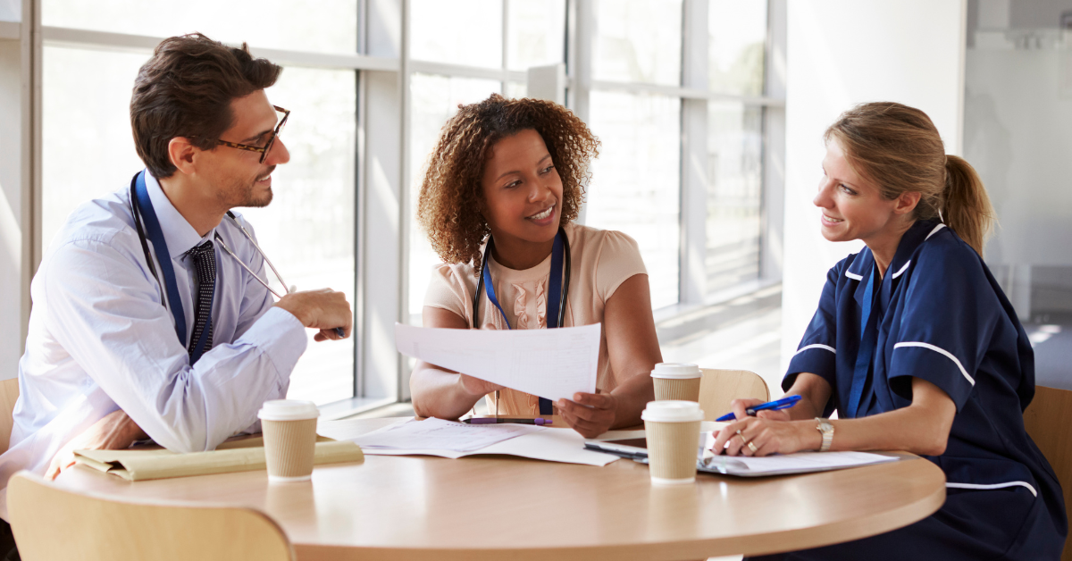 Three people, two women and one man, sit chatting at a table. The woman in the centre holds a sheet of paper, and on top of the table are two takeout coffee cups.