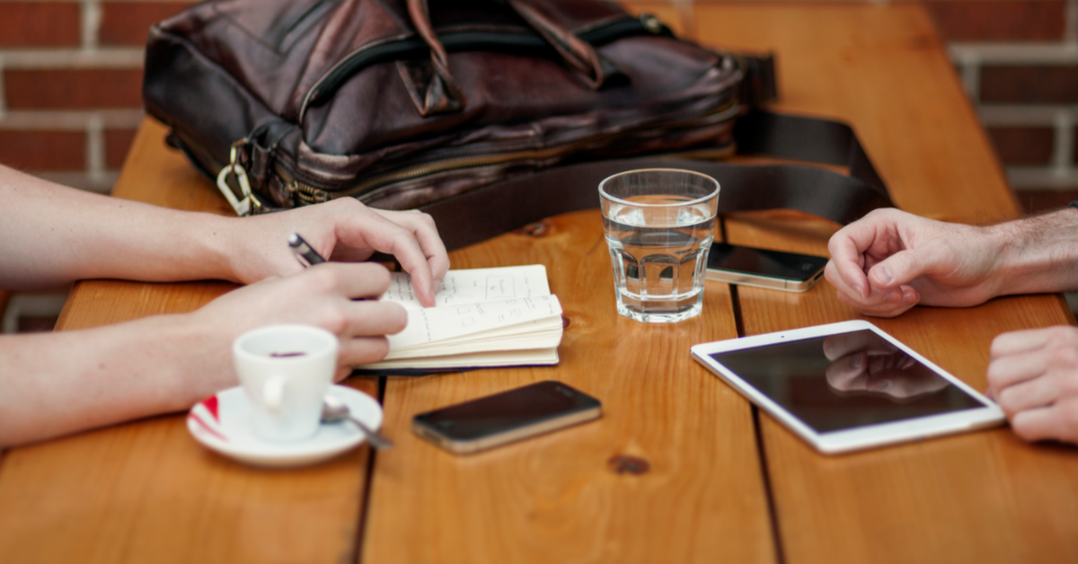 Image centres on the hands of two people sat on either side of a wooden table. In front of them are a glass of water and cup of coffee. The person on the left is writing in a notebook.