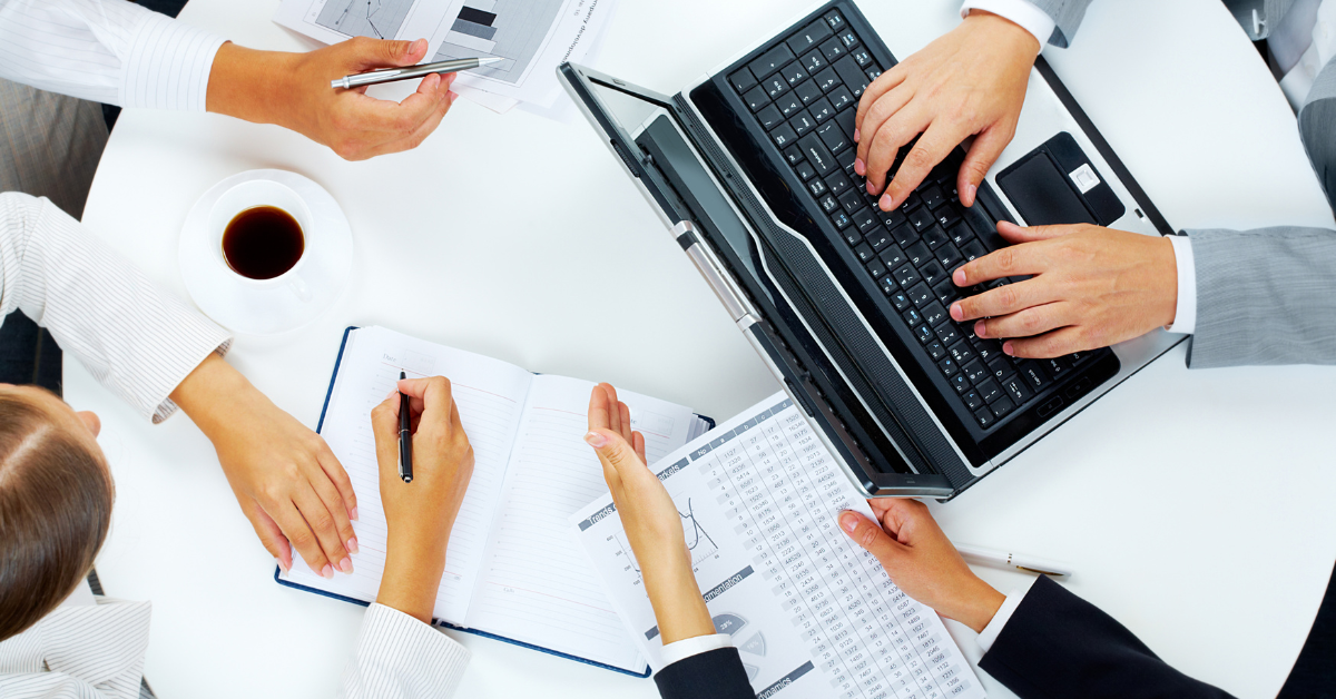 From a bird's eye view, we see the hands of four people sitting around a table. From the top right corner clockwise: one types at a laptop, one holds a page with a list of figures, one writes in a notebook, and one points at a graph with a pen.