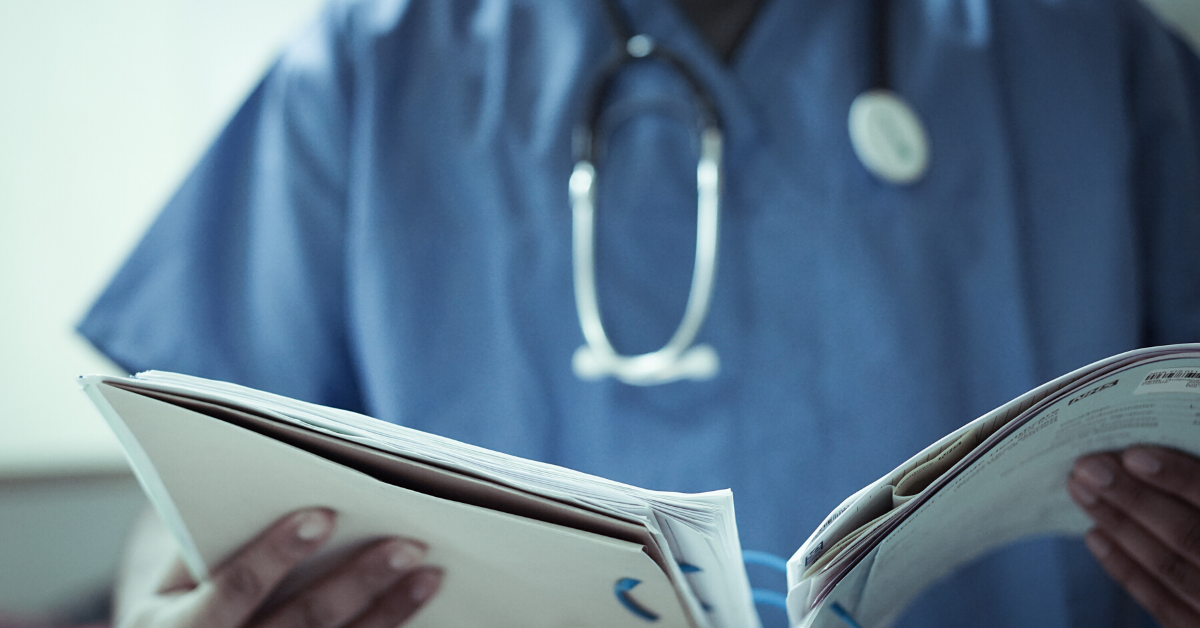 We see the torso of a medical professional dressed in blue scrubs, with a stethoscope around their neck. They are holding an open medical document in front of them.