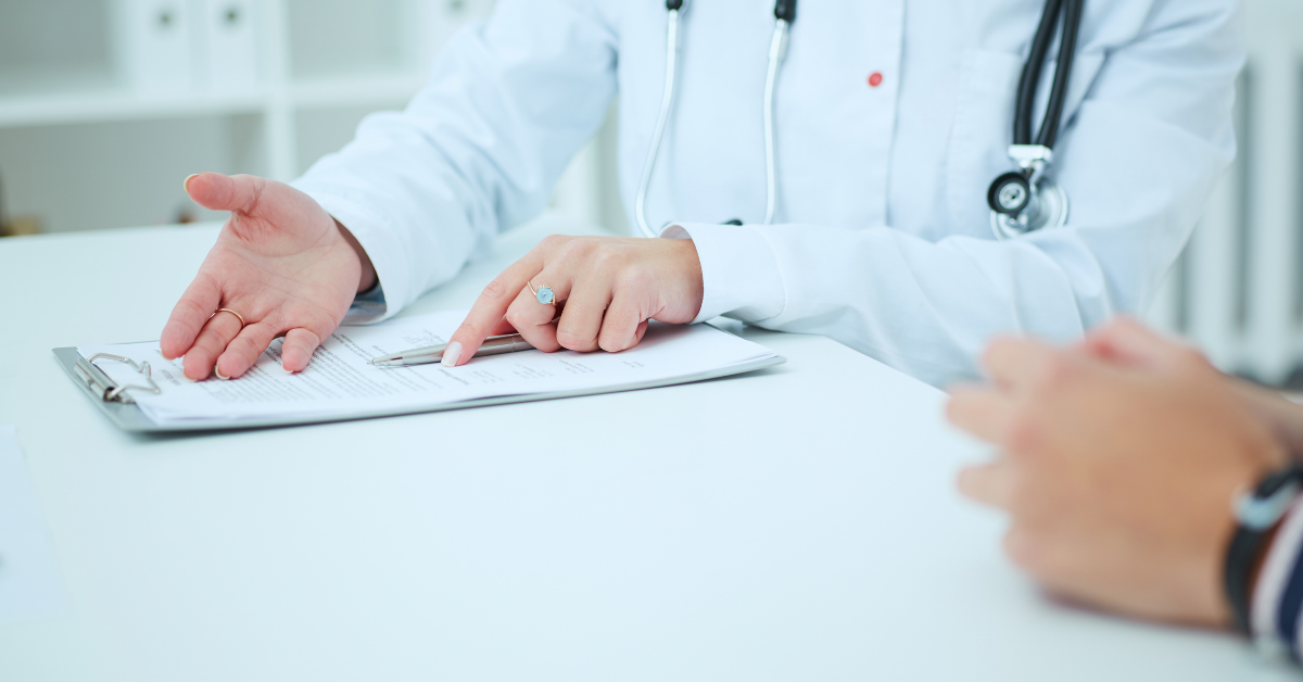 The image centres on the torso a woman sat at a white table. She is gesturing to a clipboard in front of her, and in the foreground we see the hands of the person she's speaking to.