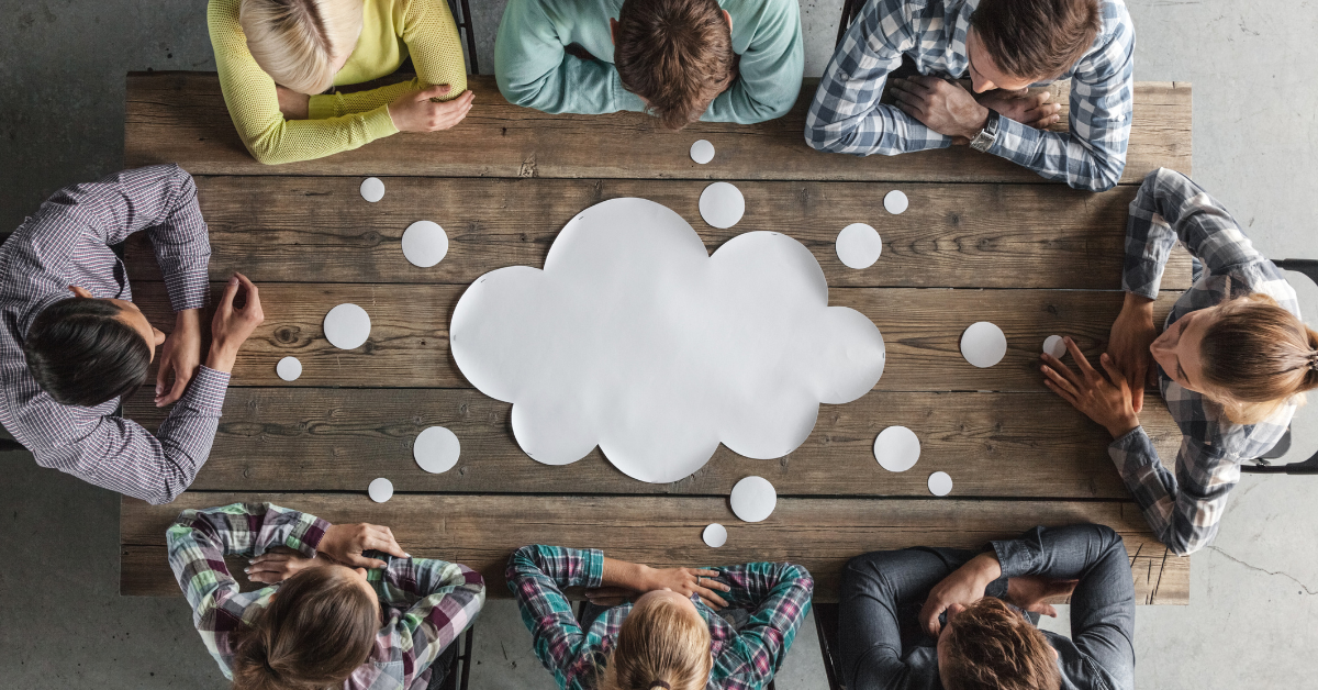 From a bird's eye view we see eight people sat around a wooden table. They all have their arms folded, and in the middle of them is a large paper thought bubble.