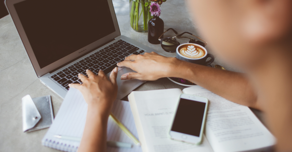 A person types on a grey laptop. By their right arm is a full mug of coffee, and in front of them is an open book with a mobile phone on top of it. By their left wrist is a spiral-bound notebook and two pens.