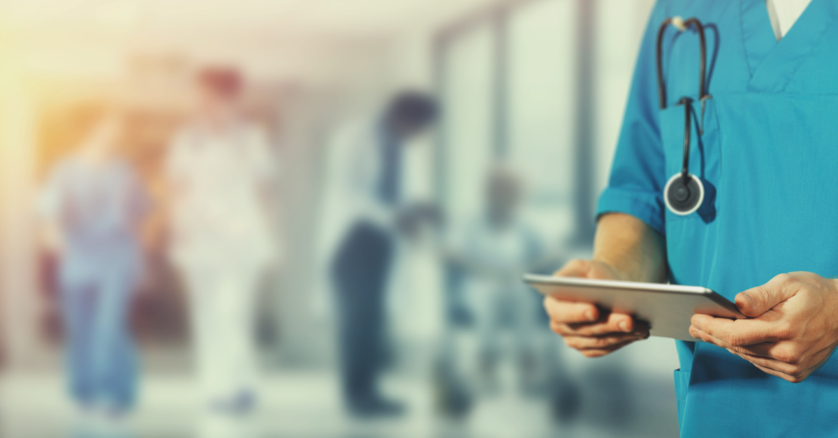 In the foreground of the photo is a medical worker, dressed in teal-coloured scrubs with a stethoscope hanging out of his pocket. He holds a tablet.