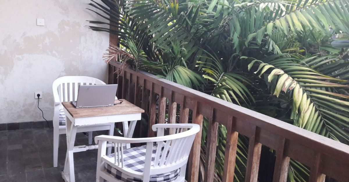 In the centre of the image is a small table, with two white wooden chairs, on a wooden balcony. On top of the table is a laptop, plugged in at the wall behind it. Behind the wooden rail at the right of the image is lush green foliage.