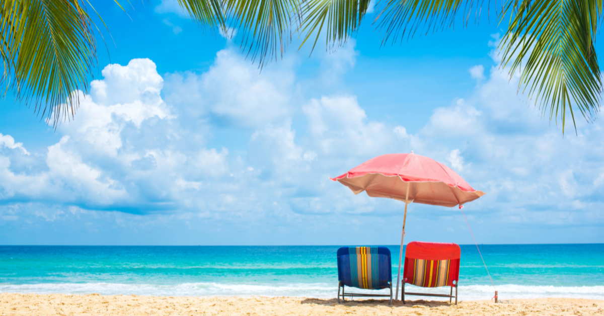 On a sandy beach, two striped deckchairs (one blue, one red) sit next to each other in front of a calm ocean and blue sky. Over them is a large pink umbrella. At the top of the image we see the leaves of palm trees.