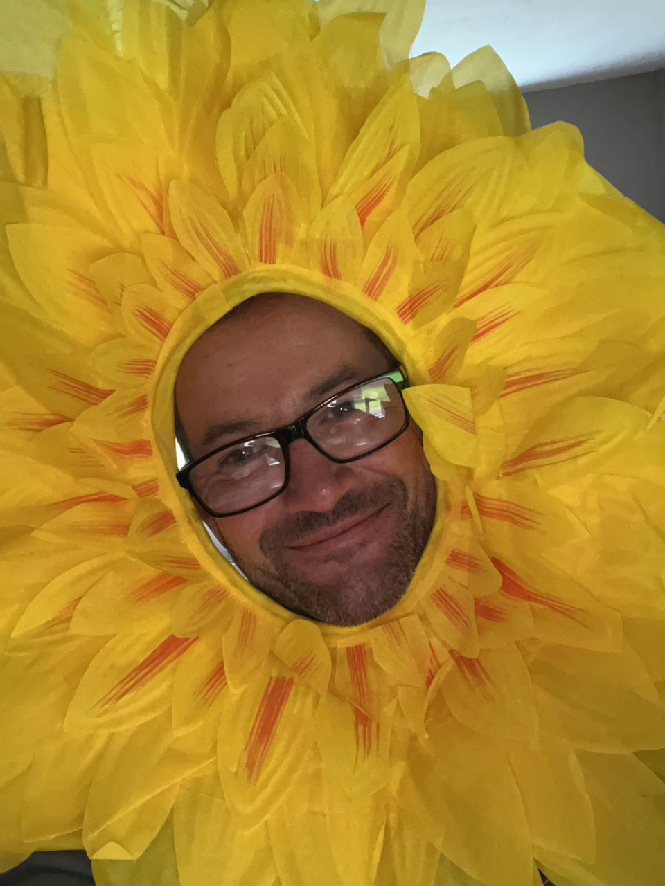 A bespectacled man, wearing a large sunflower headdress, smiles at the camera