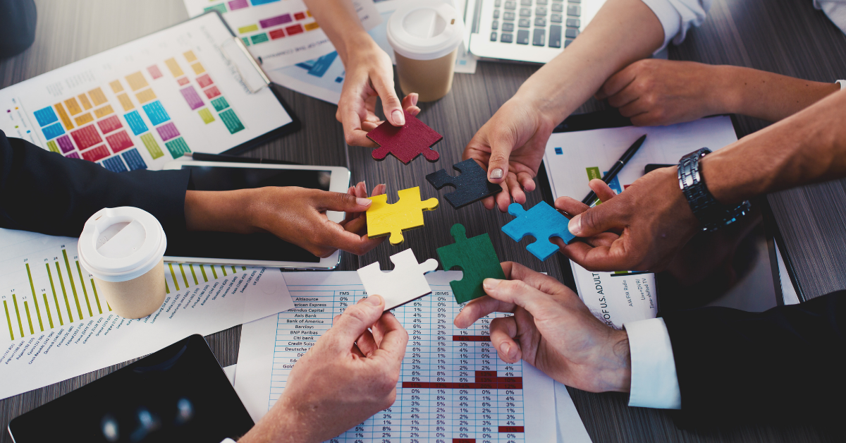 Six hands hold a different coloured jigsaw puzzle piece each, together in the centre of a table. Also on the table are various sheets of paper displaying data and figures.