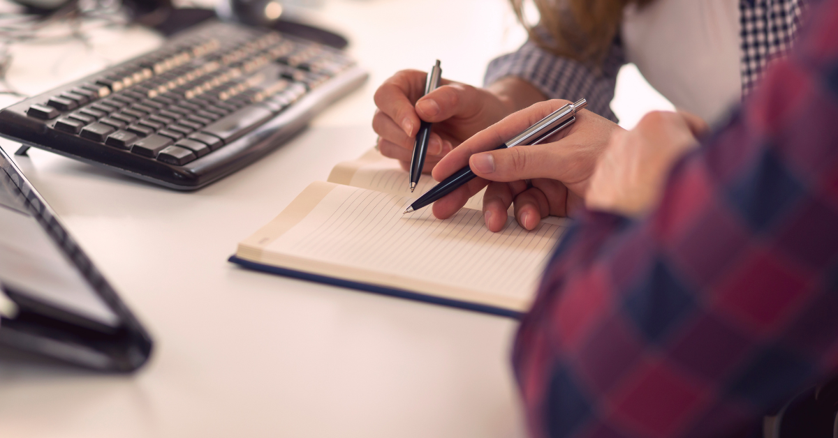 We see the hands of two people writing in a notebook on top of a desk.