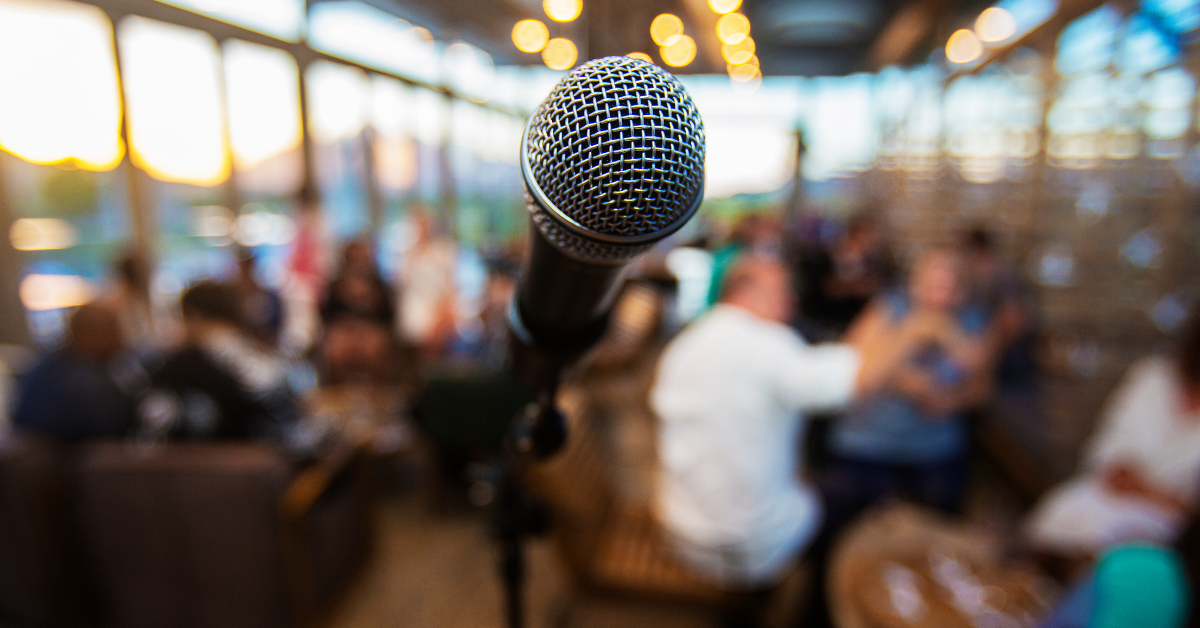 A microphone stands in the foreground. In the background, blurred slightly, is a crowd of people sat round tables.