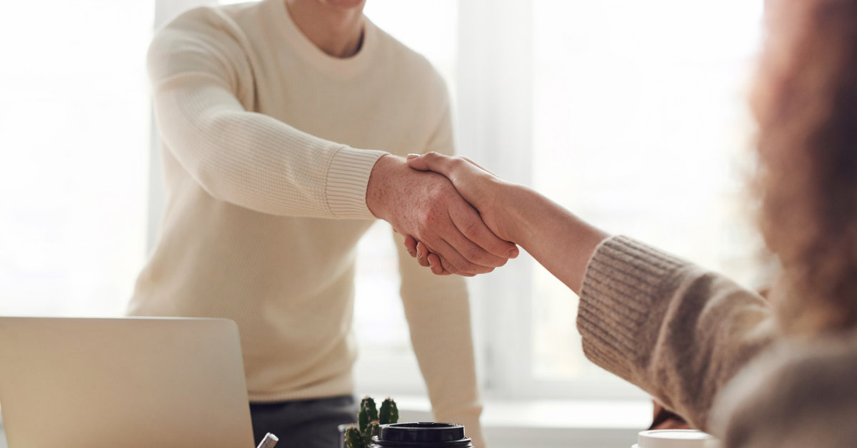 Two people, a man and a woman, shake hands over a desk. The man is stood up in the background of the image, with the woman sitting in the foreground.