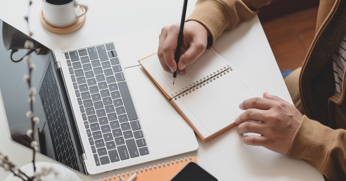 Image shows a white desk, with an open laptop. In front of it sits a person writing in a notebook - we can only see the torso and hands.