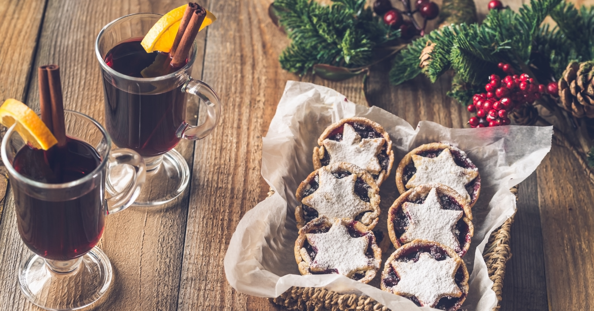 A basket of mince pies sits at the front of the image, on a wooden table. Next to the mince pies are two glasses of mulled wine, accompanied with a slice of orange and stick of cinnamon.