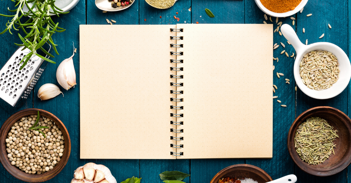 In the centre of the image is an open, blank spiral-bound notebook. Surrounding it are small bowls of various herbs and spices.