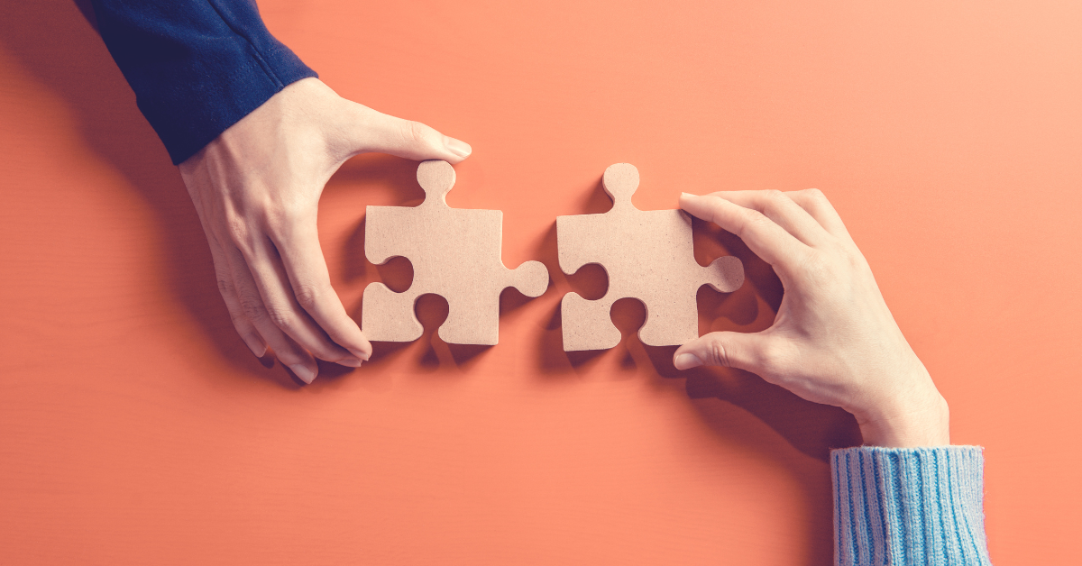 Against an orange background, we see two separate hands each holding two large, wooden puzzle pieces. They are poised as if about to fit the pieces together.