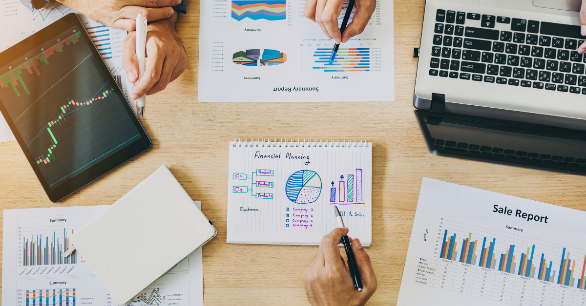 A bird's eye view shows us four people sat around a table, each holding a pen with several sheets of a report in front of them. There is a laptop in the top right corner, and a notebook with 'Financial planning' written on the page in the middle.