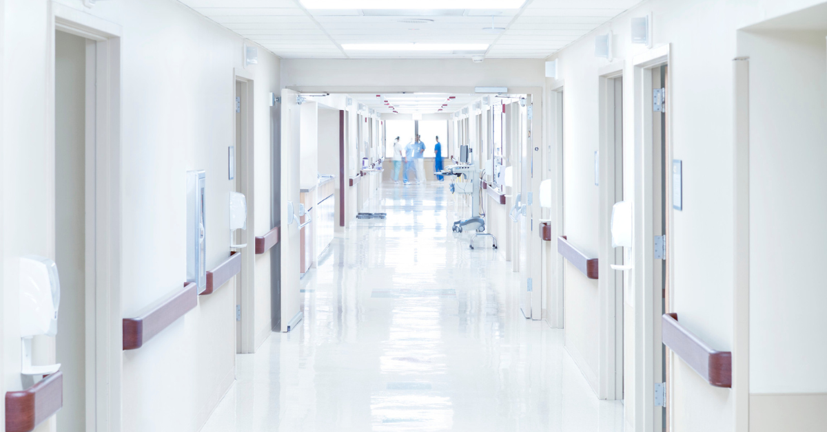 A near-empty hospital corridor stretches into the middle distance. At the very end of the corridor, a group of 4 medical staff stand chatting by a window.