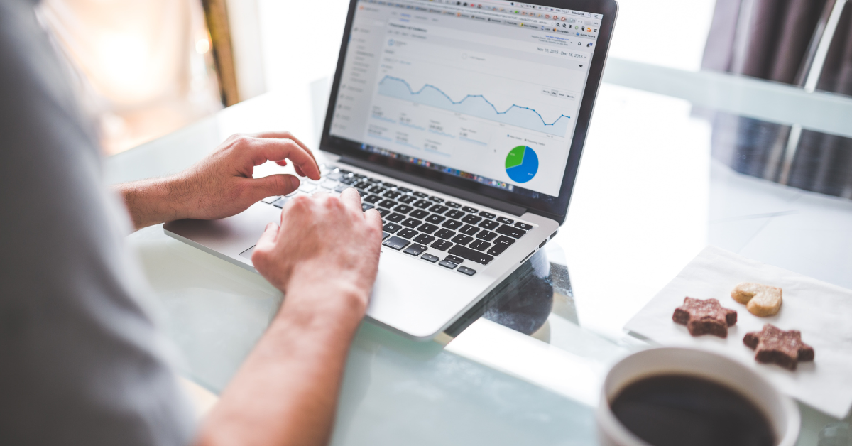 Image centres on a man's hands as he works at a laptop. On the screen is analytics data, and to the right of the laptop is a plate of biscuits and a mug of coffee.