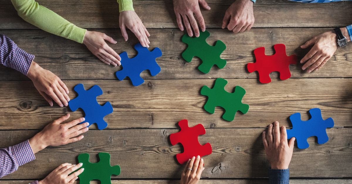 From a bird's eye view we see seven pairs of hands, each holding a large jigsaw puzzle piece across a wooden table.