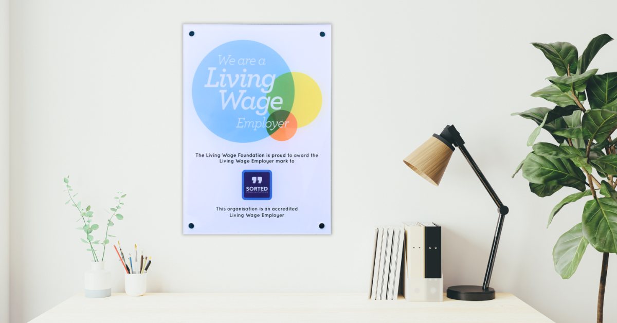 Above a tidy white desk, on a cream-coloured wall, sits a colourful Living Wage Employer certificate with the Sorted Communications logo at the bottom.