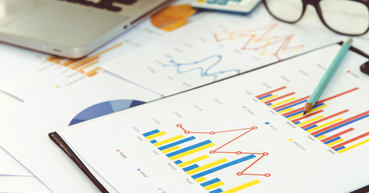 The image entirely consists of clipboards and sheets of paper displaying various multi-coloured graphs. A pale blue pencil sits across one of the graphs, towards the right side of the photo.