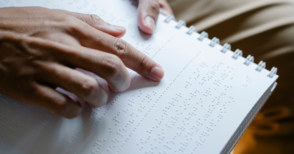 Image shows a spiral-bound book printed in braille. We see someone's hand pointing to a line of braille and reading it.