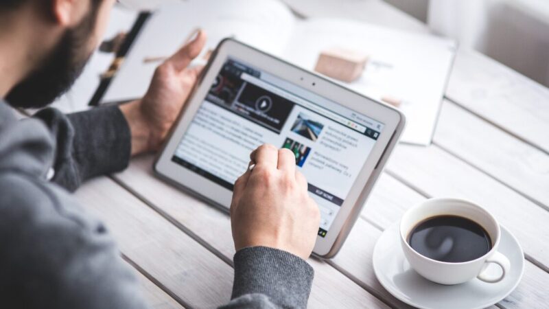 Close up of man holding a tablet in his left hand and looking at an open webpage on its screen. Next to his right hand, on the table, is a cup of black coffee.